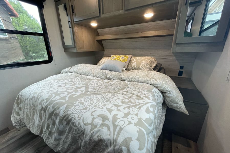Comfortable RV rental bedroom featuring a large bed with a patterned duvet, overhead cabinets, and a window overlooking greenery.