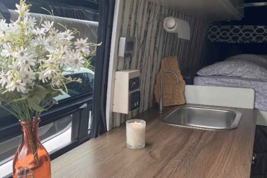 Cozy RV rental camper interior: wooden counter with sink & faucet, white cabinets, and bed. Flowers & candle add charm.