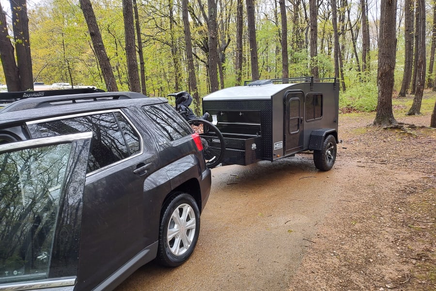 RV rental: dark SUV towing a Hiker Trailer camper & bike. Ready for adventure in a wet, lush forest setting.
