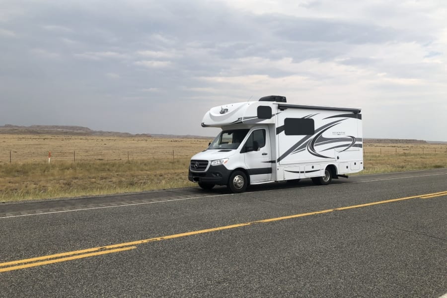 A white RV rental on a long, straight road, with vast, dry plains and distant hills under a cloudy sky. Perfect for road trips.