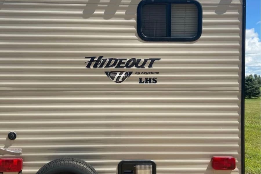 Rear view of a beige rv rental, showing its spare tire, back window, and "Hideout" decal under a blue sky.