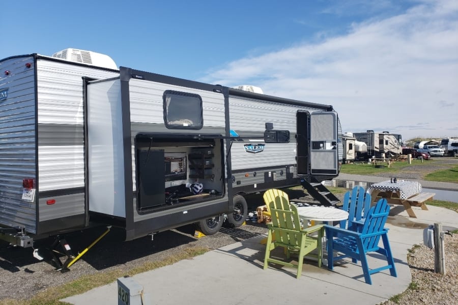 RV rental travel trailer set up at a sunny campsite with an outdoor entertainment area and patio seating.