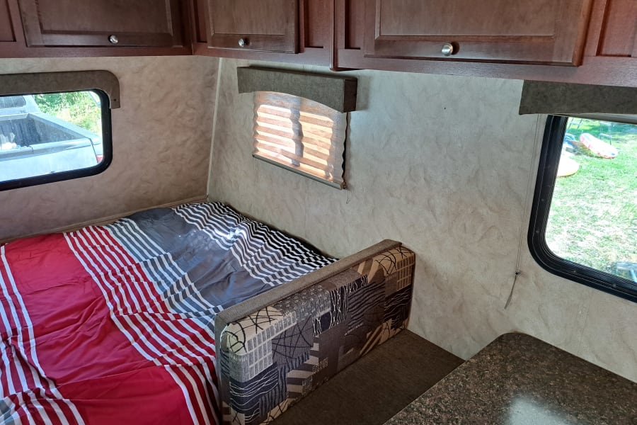 Cozy RV rental interior: striped bed, patterned seating, overhead cabinets, and outdoor views from windows.