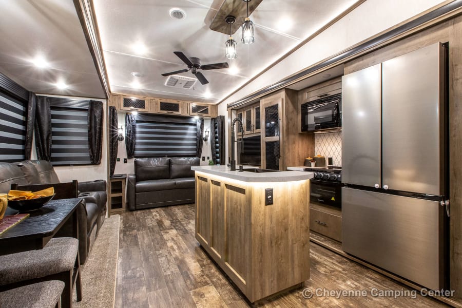 Inside this luxurious rv rental: a modern living area with sofas, a full kitchen island, stainless fridge, and warm lighting.