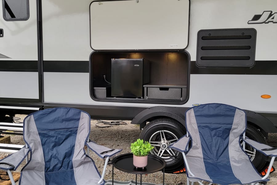 Relaxing at an RV rental campsite. Two chairs, a table, and an RV outdoor kitchen with a fridge are set up under an awning.
