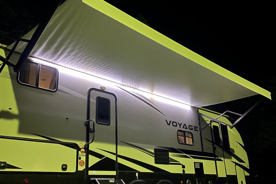 Nighttime RV rental scene. Illuminated modern RV with extended awning, steps, and mat on grass. Perfect for camping.