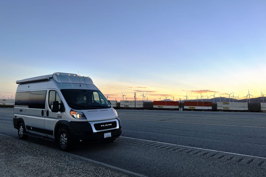 Silver RV rental camper van parked at sunset beside a long train & wind turbines.