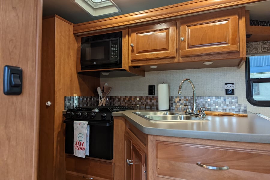 Compact RV rental kitchen with microwave, stove, double sink, and wooden cabinets.
