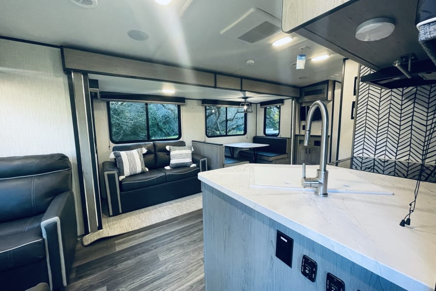 Spacious RV rental interior: living area with sofa, kitchen island with sink, and dinette. Large windows reveal outdoor scenery.