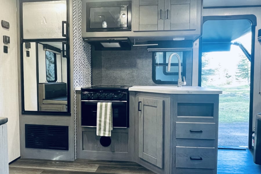 Modern RV rental kitchen with Dometic fridge, microwave, oven, sink, gray cabinets & outdoor view.