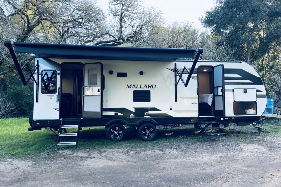 This large Mallard travel trailer, a comfortable RV rental, set up in a wooded outdoor camping area. Doors are open.