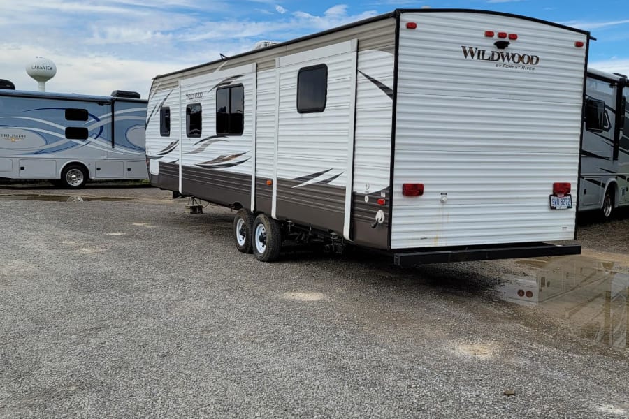 Wildwood RV rental travel trailer on a gravel lot under a blue sky. Another RV and a Lakeview water tower are visible.