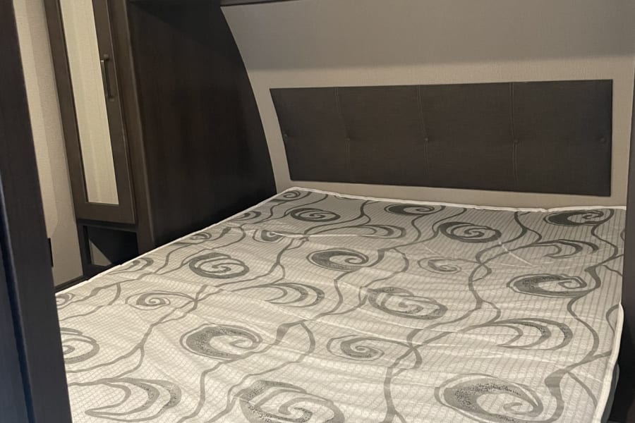 Inside an RV rental: a cozy bed with a gray swirl-patterned mattress and dark upholstered headboard, flanked by dark cabinetry.