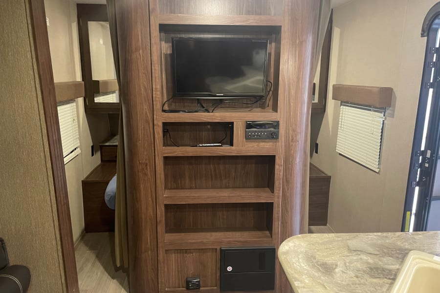 RV rental interior: A central wooden entertainment unit with TV, stereo, and shelving, adjacent to a sleeping area and kitchen space.