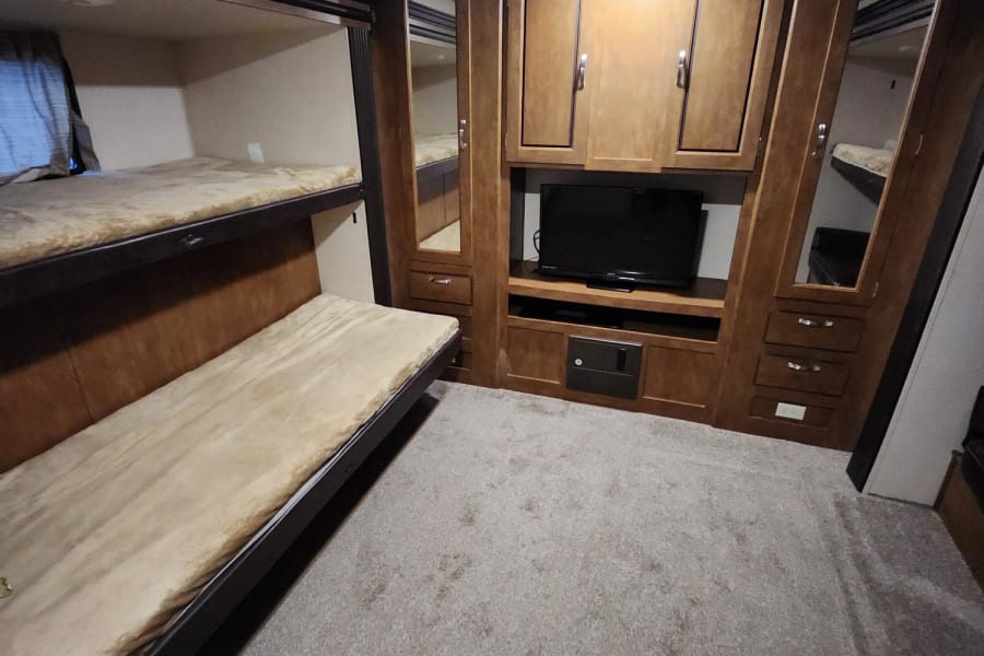 Inside an RV rental: a bedroom with a dark wood cabinet unit, TV nook, mirrors, and a bed with lift-up storage compartments. Gray carpet.
