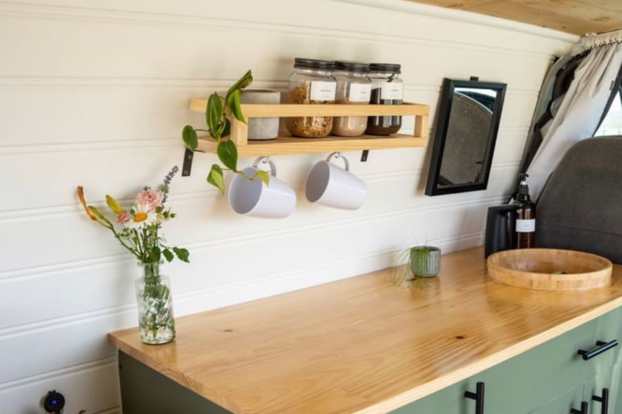 Inside an RV rental: a bright, organized kitchen with a wooden shelf, jars, hanging mugs, and counter.