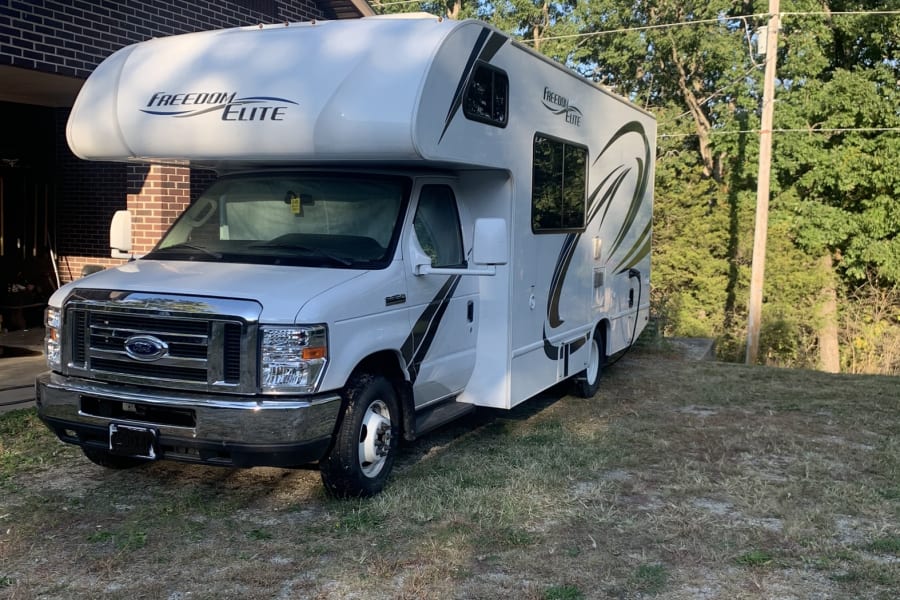 A white Freedom Elite Class C RV rental is parked on gravel next to a brick house, under a blue sky with trees.