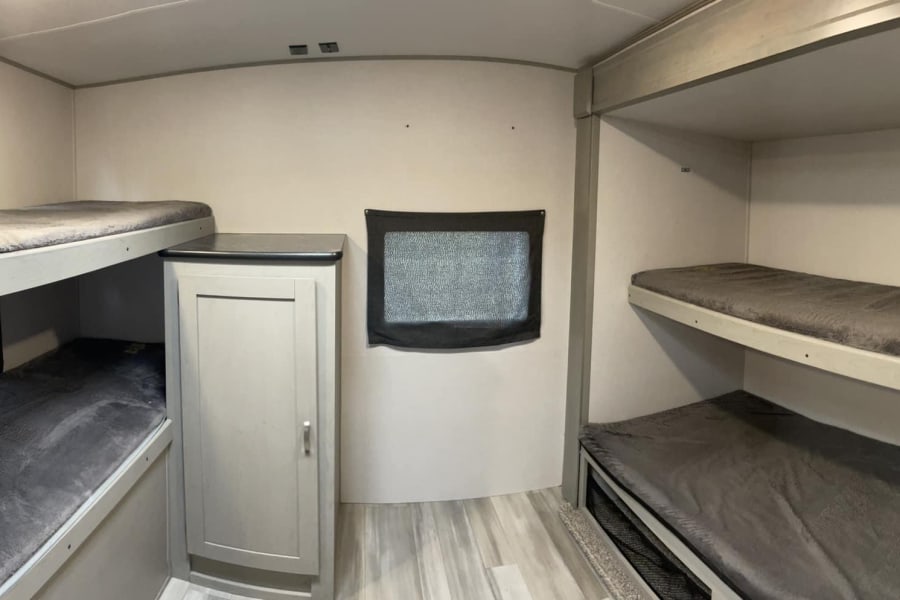 Inside an RV rental, two sets of bunk beds with grey bedding. A small cabinet and covered window are visible, offering family sleeping space.