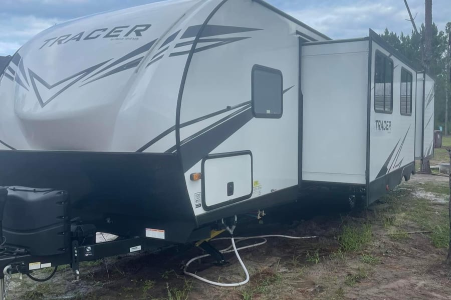 This Tracer RV rental is extended at a peaceful campsite with pine trees under a cloudy sky. Ready for adventure!