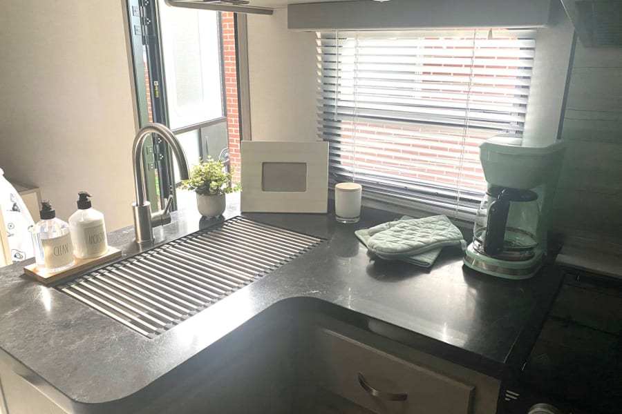 Cozy RV rental kitchen: dark counters, a stainless steel sink with drying rack, light green coffee maker, and a window.
