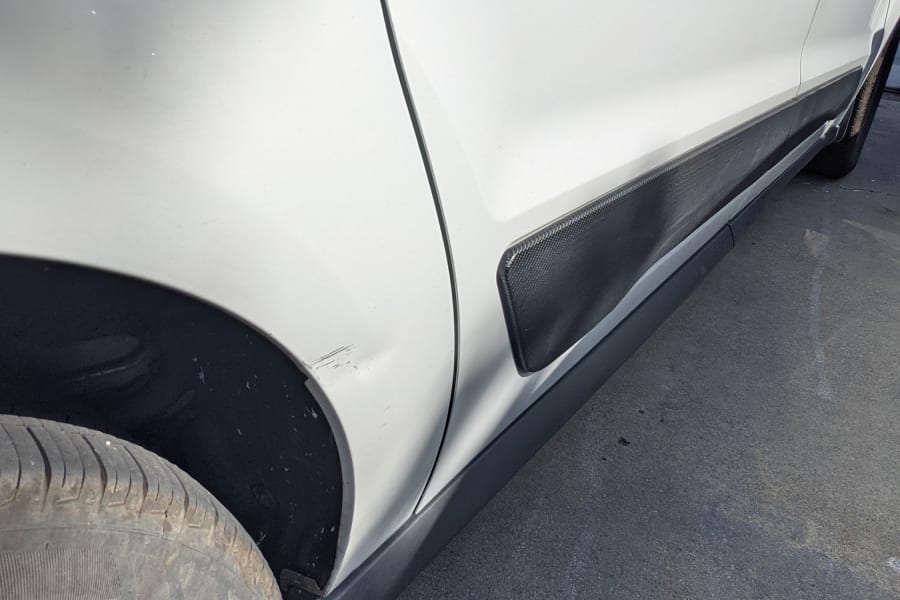 White RV rental van with a noticeable side body dent and scuff marks above the front tire.