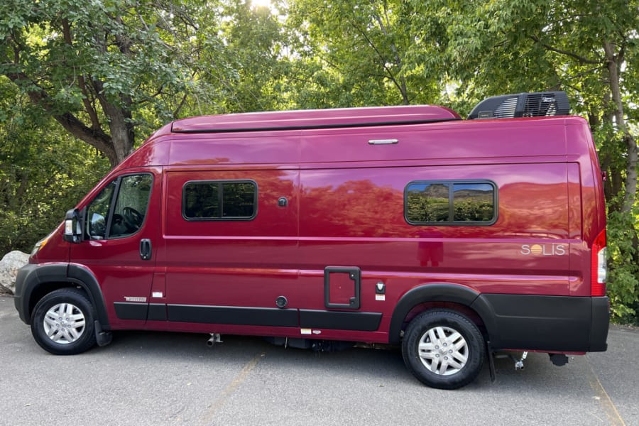 This maroon Winnebago Solis campervan is ready for your next rv rental adventure, parked among green trees.