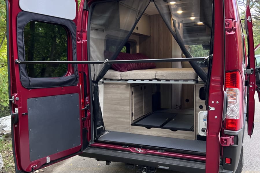 Inside an RV rental, the open rear of this red camper van shows a raised bed, mesh screen, and ample storage.
