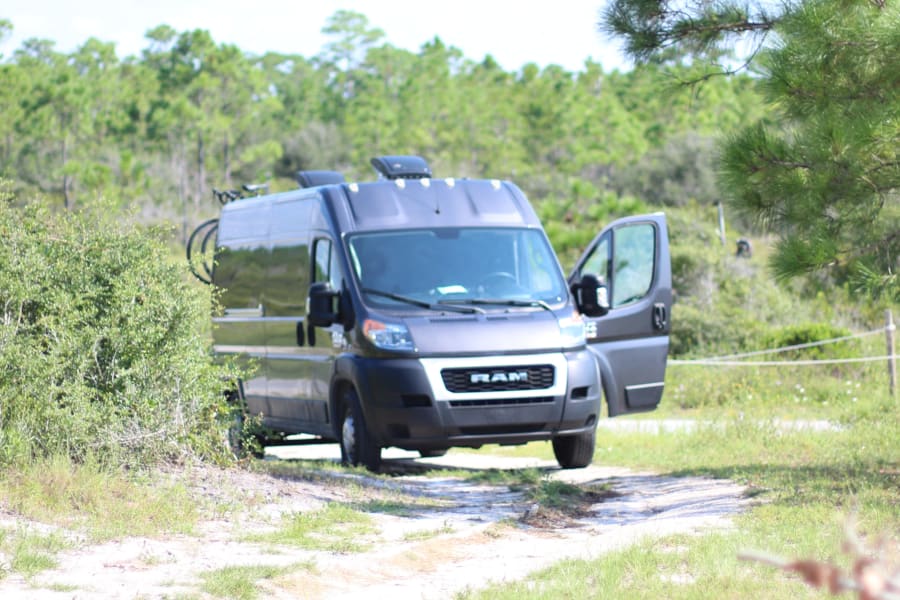 Grey RAM ProMaster RV rental van, door open, parked on a path by green foliage, ready for outdoor adventure.