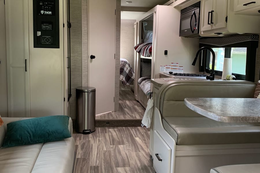 This RV rental interior offers a modern living space with light furnishings, kitchen, dining area, and rear bunk beds.