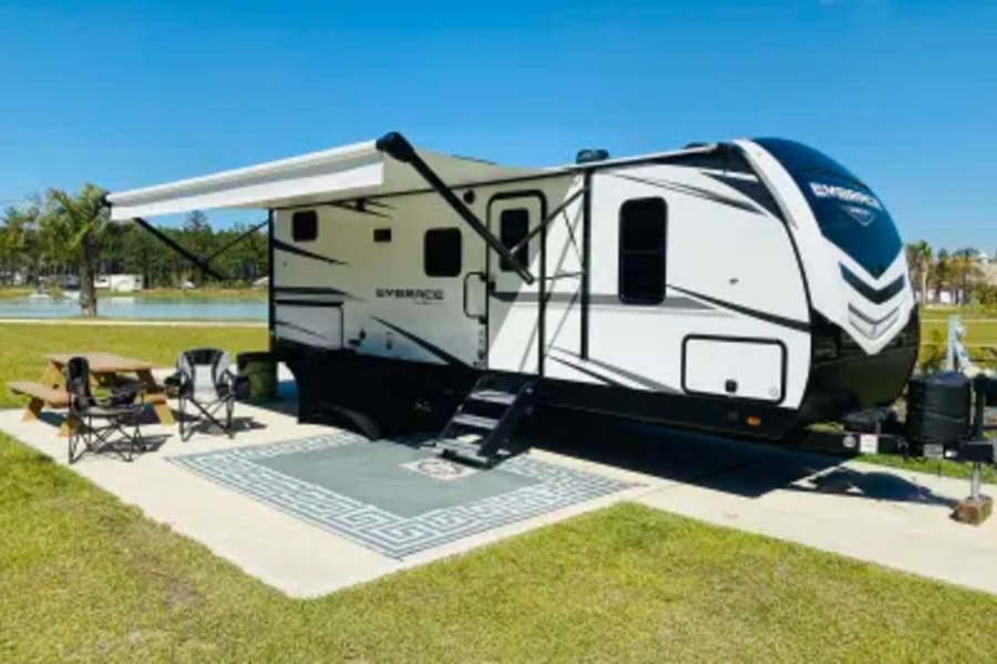 Bright day at an RV rental campsite. White RV with awning, outdoor rug, picnic table & chairs on concrete pad.