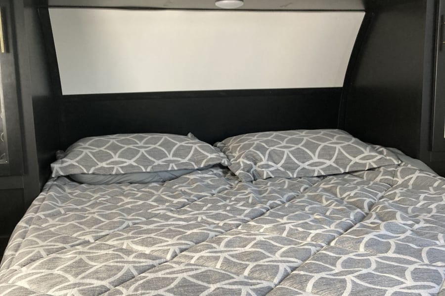 Comfortable RV rental bed featuring gray patterned bedding and dark overhead storage cabinets.