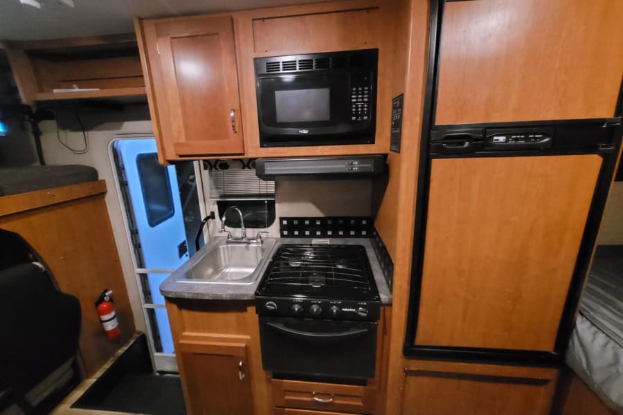 Inside this RV rental: a compact kitchen with stove, microwave, sink, and wood-paneled fridge. Fire extinguisher near exit.