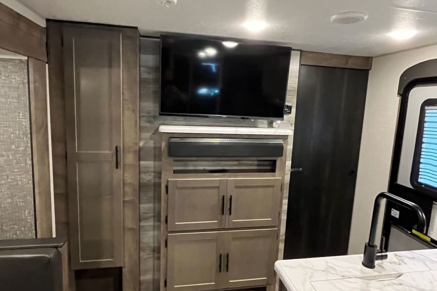 Inside a modern RV rental, a central entertainment area with TV/soundbar, full kitchen island, and dining booth creates a functional living space.