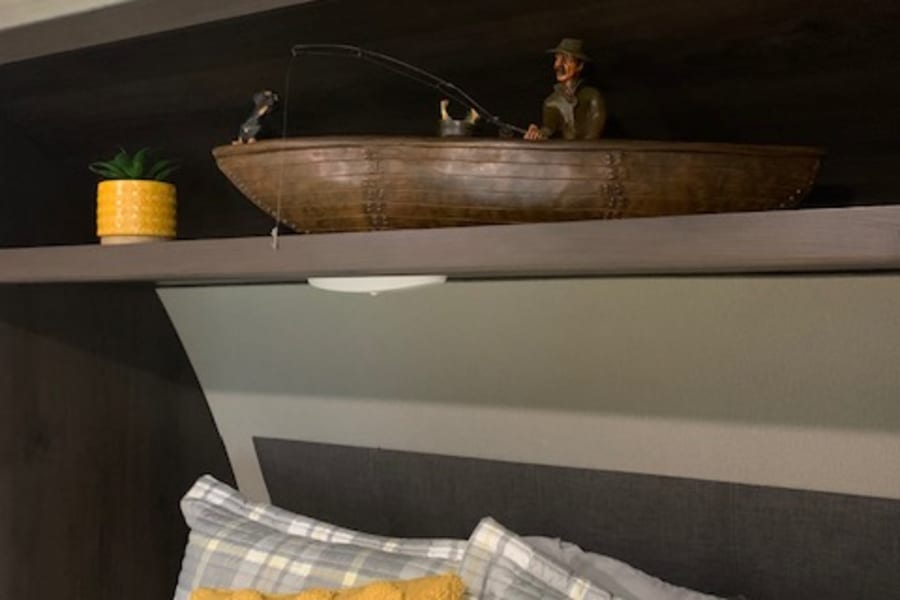RV rental bed area with cozy plaid bedding, yellow throw pillow, and a unique shelf displaying a fishing boat figurine.