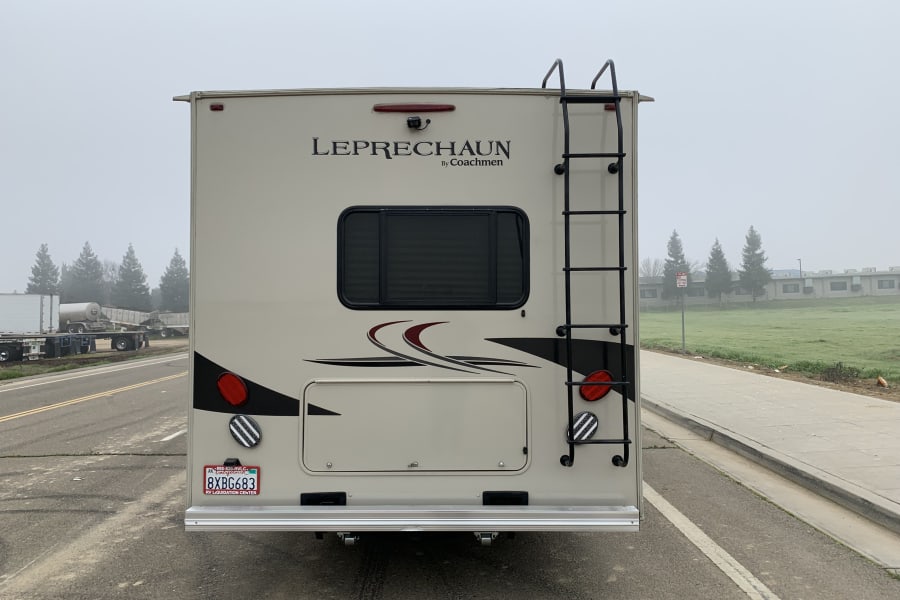 Rear of a beige Coachmen Leprechaun RV rental motorhome. Features a ladder, camera, and window, parked on a misty day.