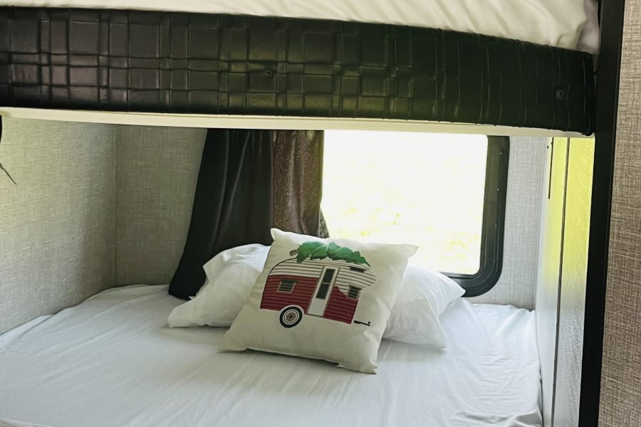 An RV rental's interior shows cozy bunk beds with white bedding & a decorative camper pillow by a bright window.