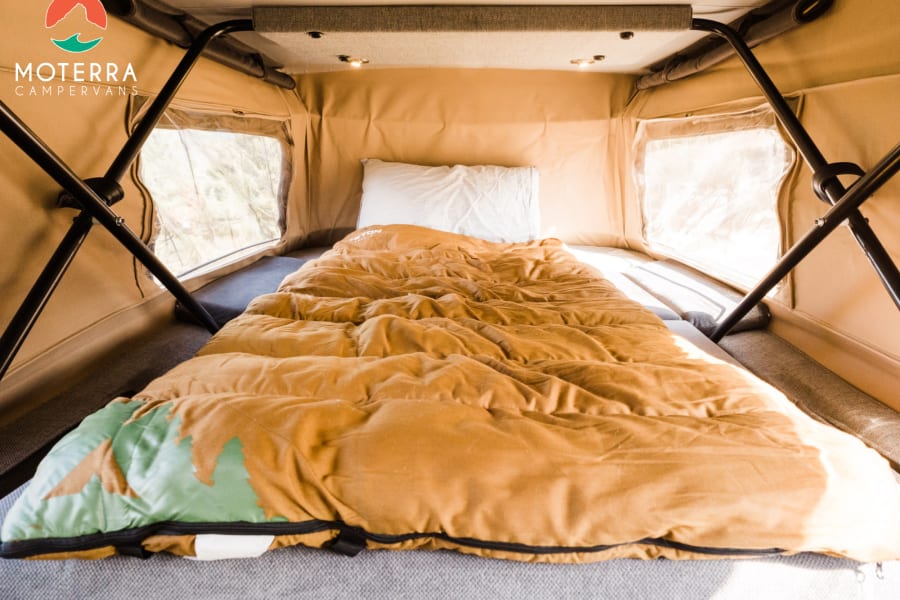 RV rental's cozy pop-top sleeping area. A bed with a brown sleeping bag and white pillow, bathed in natural light.
