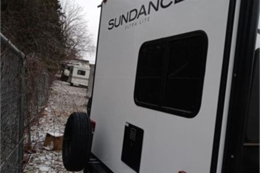 Rear view of a white Sundance Ultra Lite RV rental, featuring a mounted spare tire and back window, parked on gravel.