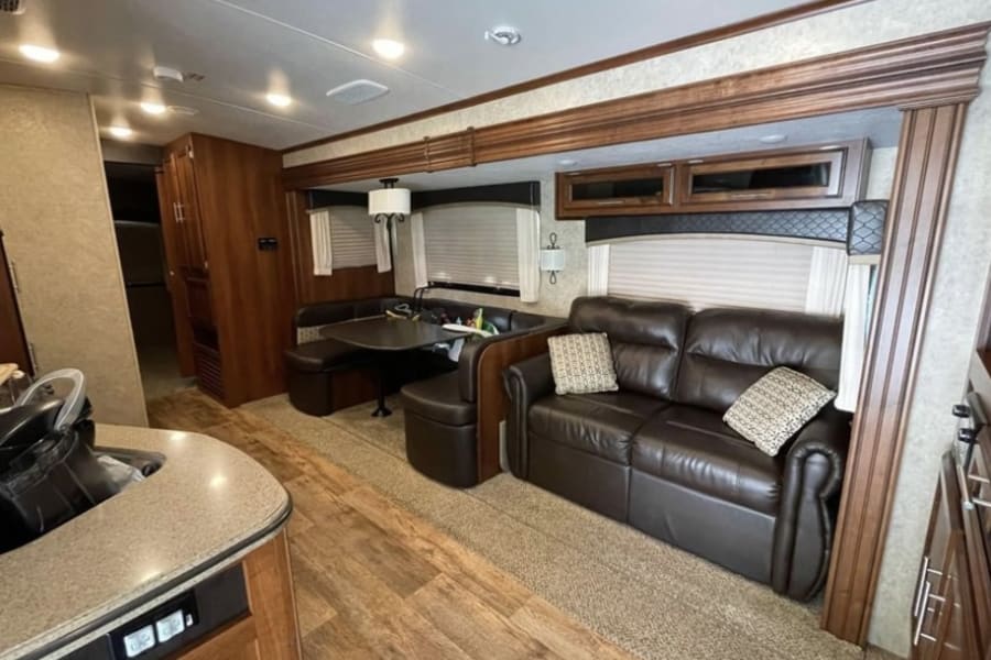 Bright RV rental interior showing a brown leather sofa, U-shaped dinette, and kitchen. Comfortable living space.