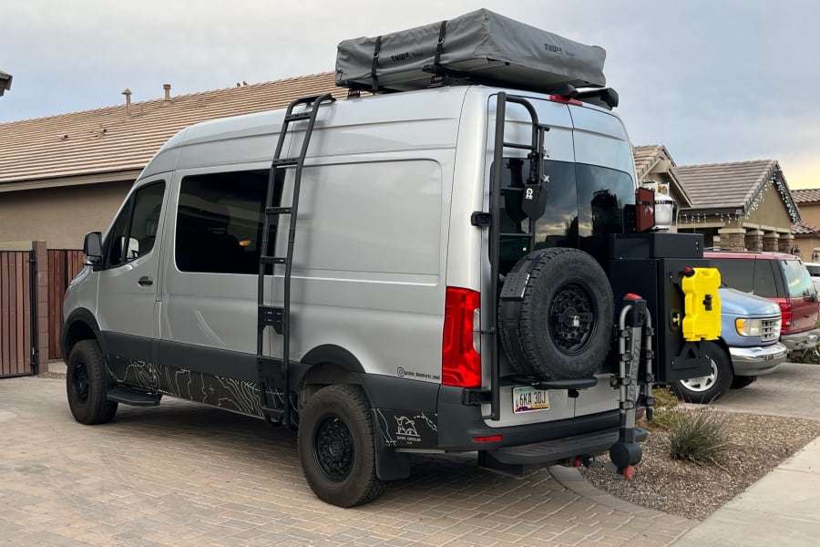 This custom Sprinter adventure van, perfect for an rv rental, features a roof tent, ladder, and rear utility setup.