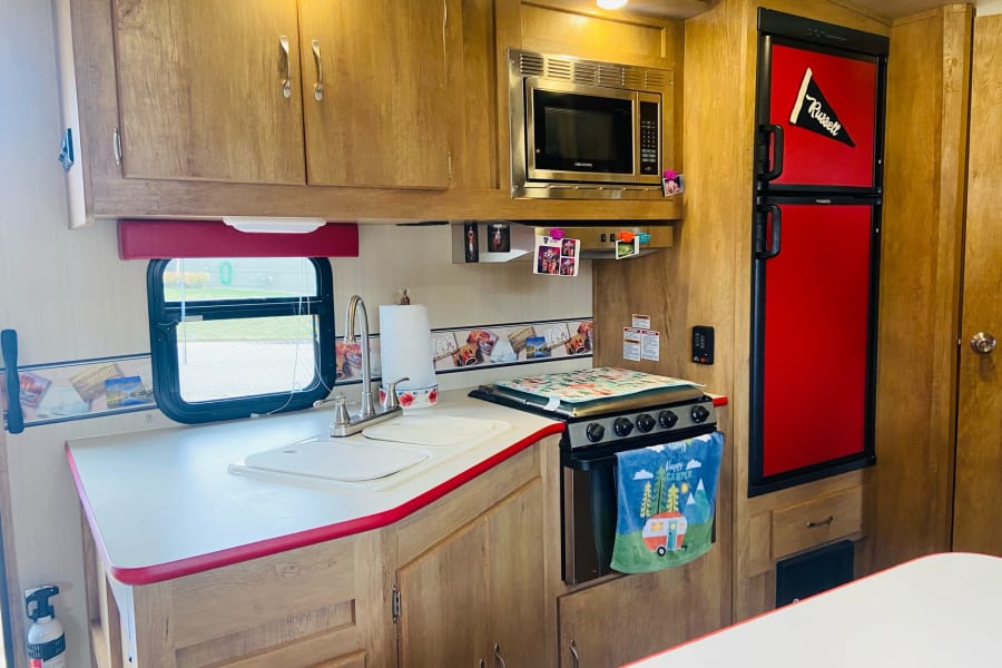 Compact RV kitchen interior with wood cabinets, red accents, microwave, fridge, sink, and stove, perfect for an rv rental.
