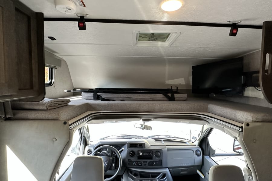 Explore this RV rental's interior: spacious overhead bunk with a safety net, driver's cabin, and mounted TV for comfortable travel.