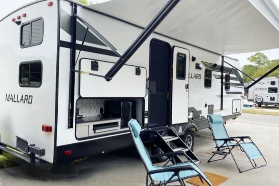 White RV rental travel trailer with an extended awning, open outdoor kitchen, and two blue lounge chairs.