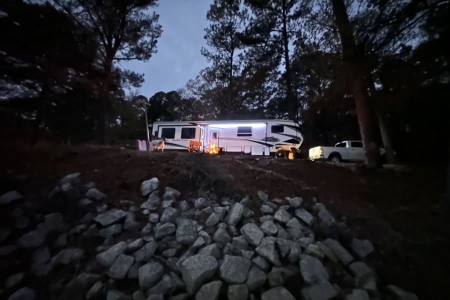 Illuminated RV rental, campfire, and dog parked in a wooded area at dusk, with a truck nearby. Ideal for a night of camping.