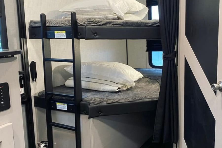 Cozy RV rental interior: two bunks with bedding, a ladder, and a fire extinguisher below. Ideal for family road trips.