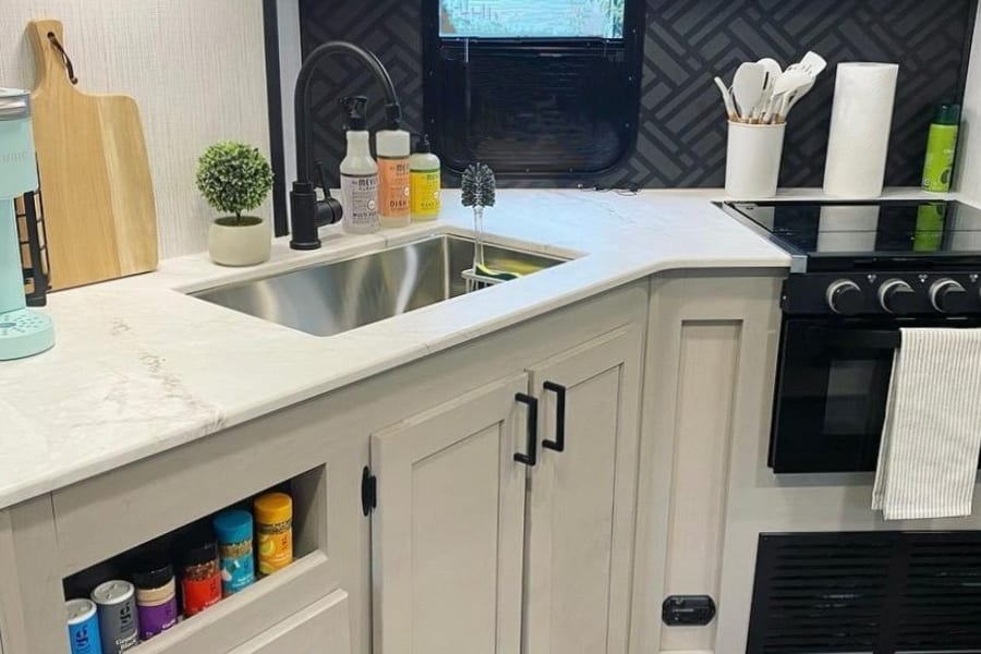 Modern RV kitchen with light countertops, black appliances, organized sink area, and stylish accent wall. Ideal for an RV rental.