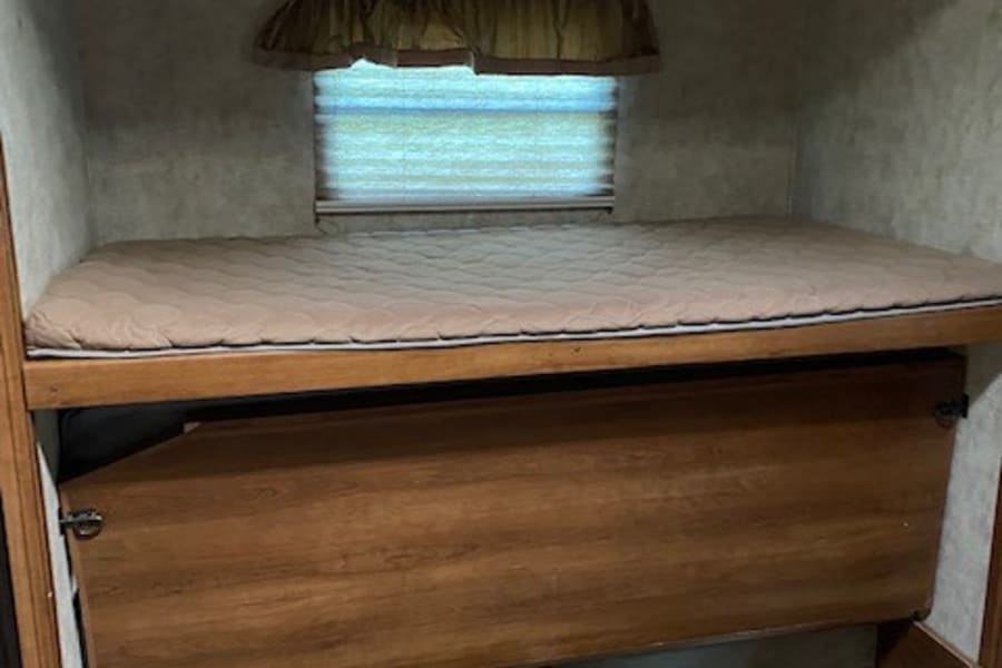 RV rental interior showing a bunk bed with mattress, window, and a lower wooden panel possibly for storage.