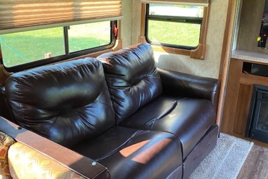 Spacious RV rental living area featuring a comfortable brown leather sofa, windows with blinds, and ample storage cabinets.