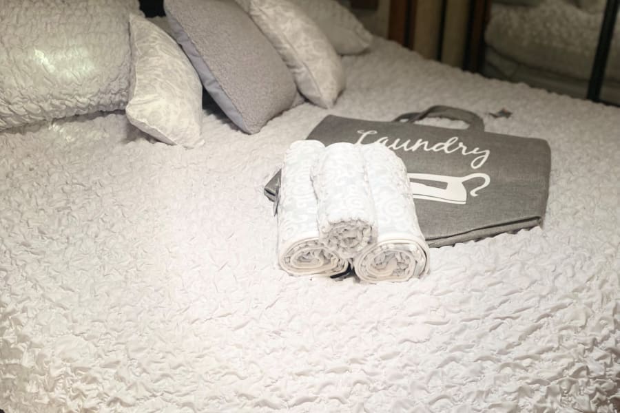 Cozy RV rental bedroom with white textured bedding, assorted pillows, rolled towels, and a "LAUNDRY" bag.