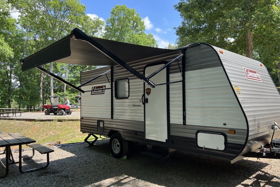 Travel trailer RV rental with extended awning at a sunny gravel campsite, surrounded by trees.